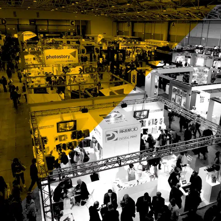 Vue plongeante d’un salon professionnel animé avec stands, signalétique et visiteurs, N&B rehaussé de jaune — événements & goodies Yescom à Pontivy.