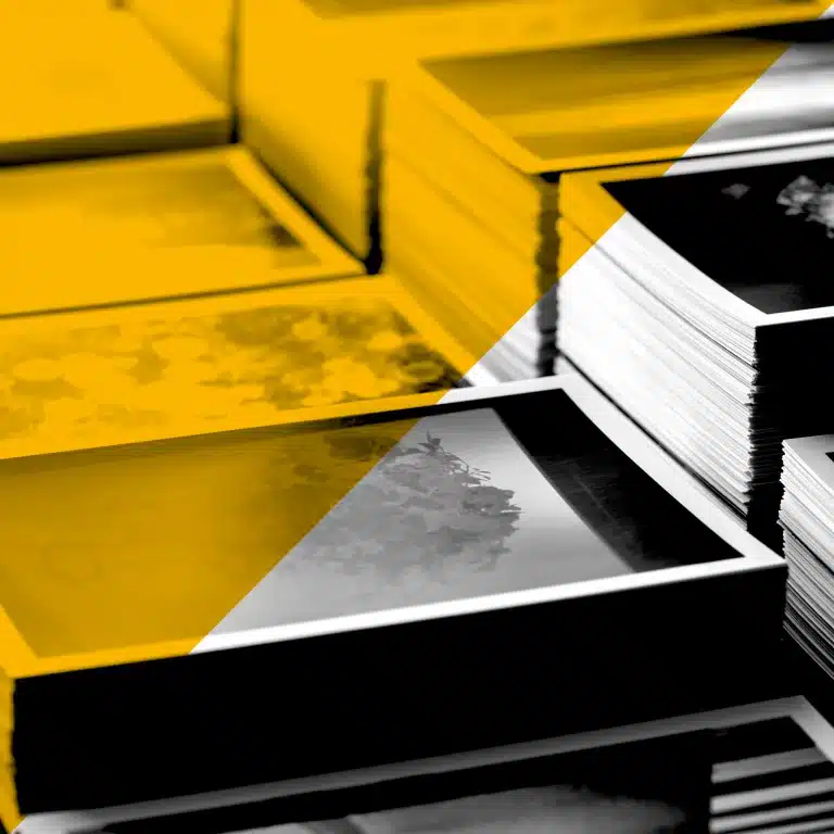 Gros plan sur piles de tirages, photos et brochures sur table d’impression, N&B avec aplats jaunes — services print Yescom à Pontivy.