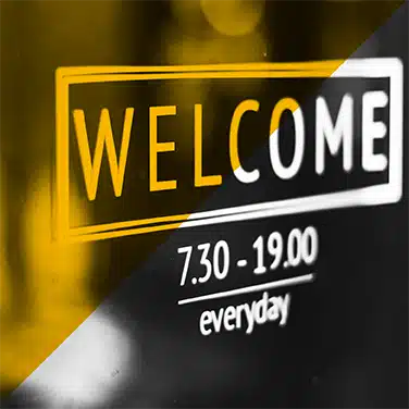 Image noir & blanc avec un aplat jaune transparent. Stickers technologique avec projection lumineuse vitrine avec inscription « WELCOME », horaires d’ouverture en dessous.